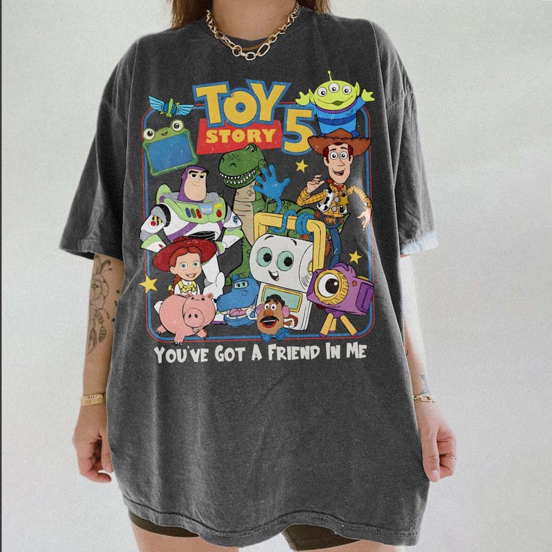 Pu&ograve; includere: T-shirt grigio scuro con una grafica colorata dei personaggi di "Toy Story 5". Il design include Buzz Lightyear, Woody e altri personaggi, con la scritta "YOU'VE GOT A FRIEND IN ME".