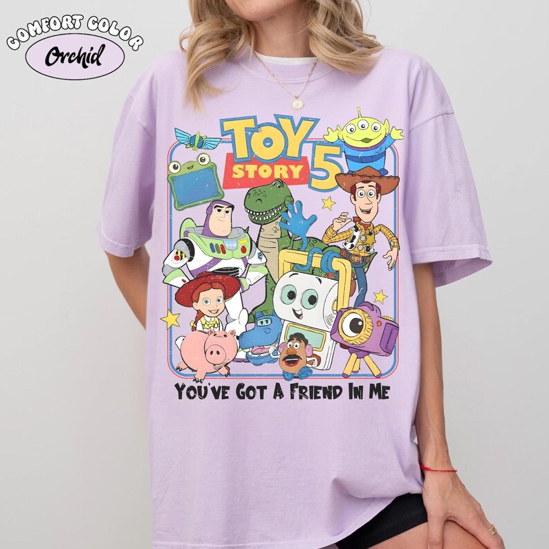 Pu&ograve; includere: Una maglietta Comfort Colors lilla chiaro con una grafica colorata di Toy Story. Il design presenta personaggi come Buzz Lightyear e Woody, con il testo "Toy Story 5" e la frase "You've Got a Friend in Me."