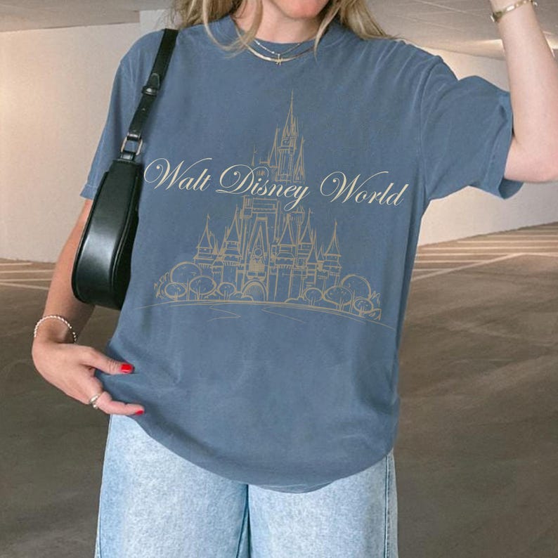 K&ouml;nnte beinhalten: Ein blaues T-Shirt mit einer goldenen Illustration eines Schlosses und den Worten "Walt Disney World". Das T-Shirt hat kurze &Auml;rmel und einen Rundhalsausschnitt. Das Design ist auf der Vorderseite des T-Shirts zentriert.