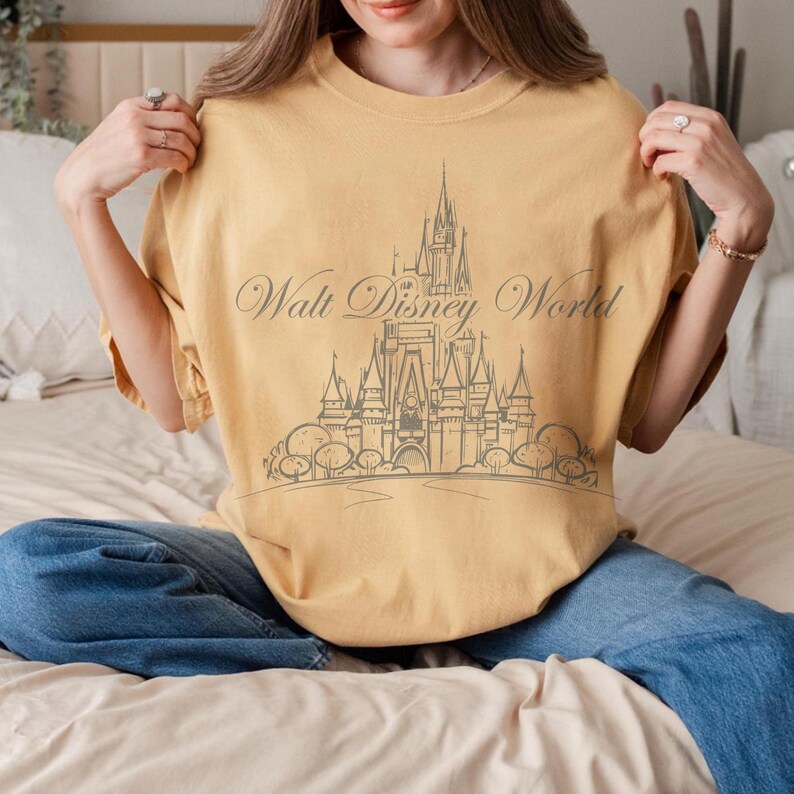 K&ouml;nnte beinhalten: Senfgelbes T-Shirt mit einer grauen Grafik eines Schlosses und dem Schriftzug "Walt Disney World". Das Shirt hat eine lockere Passform und kurze &Auml;rmel. Die Person tr&auml;gt eine blaue Jeans.