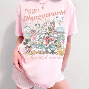 Peut inclure: T-shirt rose clair avec un motif de personnages et de monuments de Disney World. Le texte "FLORIDA Est. 1971" et "Disneyworld" sont imprim&eacute;s au-dessus du motif. La phrase "the happiest place on earth" est imprim&eacute;e en dessous.