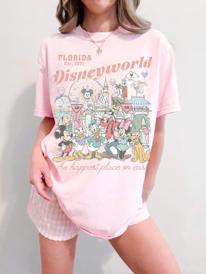 Peut inclure: T-shirt rose clair avec un motif de personnages et de monuments de Disney World. Le texte "FLORIDA Est. 1971" et "Disneyworld" sont imprim&eacute;s au-dessus du motif. La phrase "the happiest place on earth" est imprim&eacute;e en dessous.