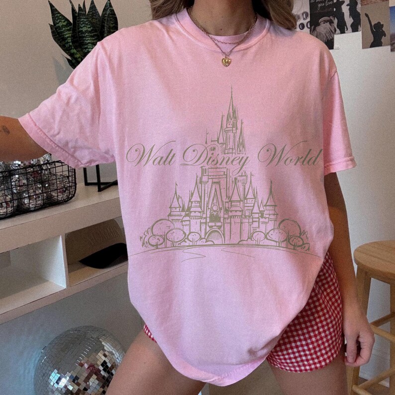 K&ouml;nnte beinhalten: Hellrosa T-Shirt mit goldfarbener Illustration von Cinderella's Schloss und dem Schriftzug "Walt Disney World". Das Shirt hat kurze &Auml;rmel und einen Rundhalsausschnitt.