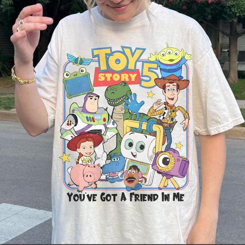Pu&ograve; includere: T-shirt color crema con una grafica colorata dei personaggi del film "Toy Story 5". Il design include Buzz Lightyear, Woody e altri personaggi, con la scritta "You've Got a Friend in Me".