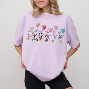 Może przedstawiać: Jasnofioletowa koszulka z kolorową grafiką postaci Disneya trzymających balony w kształcie Myszki Miki. Postacie to Pluto, Goofy, Myszka Miki, Myszka Minnie, Kaczka Daisy i Kaczor Donald. Koszulka ma luźny kr&oacute;j.