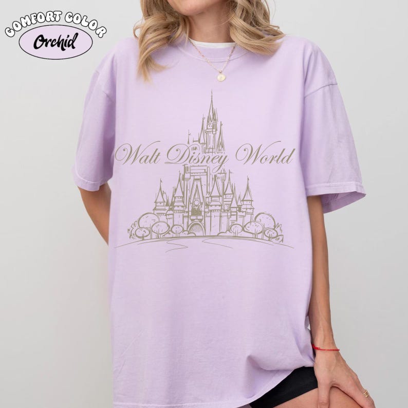 K&ouml;nnte beinhalten: Ein helllila T-Shirt mit einer Grafik von Cinderella's Schloss und den Worten "Walt Disney World" in einer Schreibschrift. Das Shirt hat auch die Worte "Comfort Color" und "Orchid" in einem Kreis.