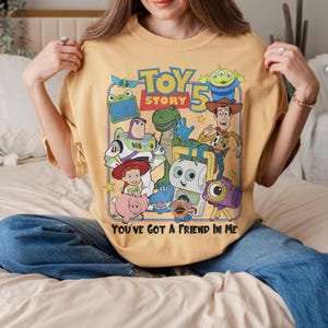 Pu&ograve; includere: T-shirt giallo senape con una grafica colorata dei personaggi di "Toy Story 5". Il design include Woody, Buzz Lightyear e altri personaggi. Il testo "TOY STORY 5" e "YOU'VE GOT A FRIEND IN ME" sono anch'essi visualizzati.