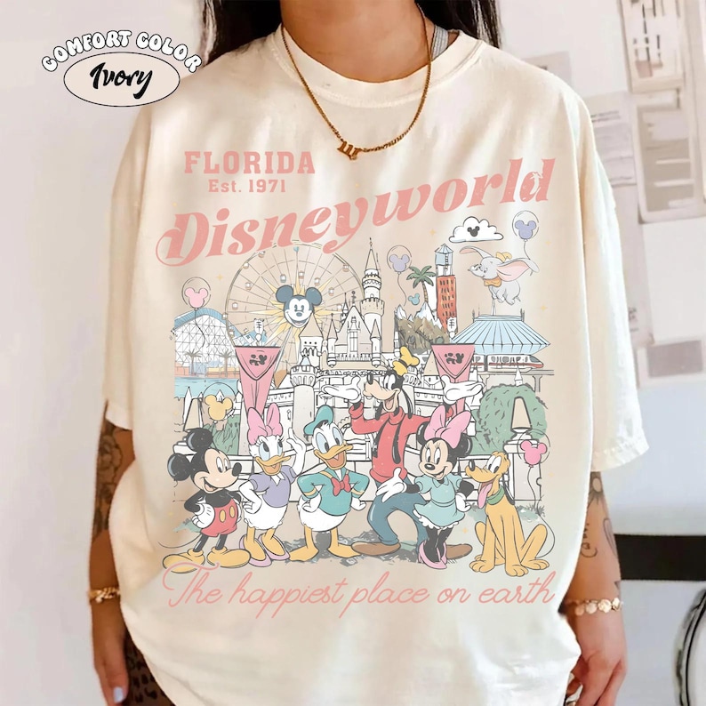 Peut inclure: T-shirt ivoire Comfort Color avec un motif de Disney World. Le design comprend le texte "FLORIDA Est. 1971 Disneyworld" et la phrase "The happiest place on earth". Mickey, Minnie, Donald et Dingo sont &eacute;galement repr&eacute;sent&eacute;s.
