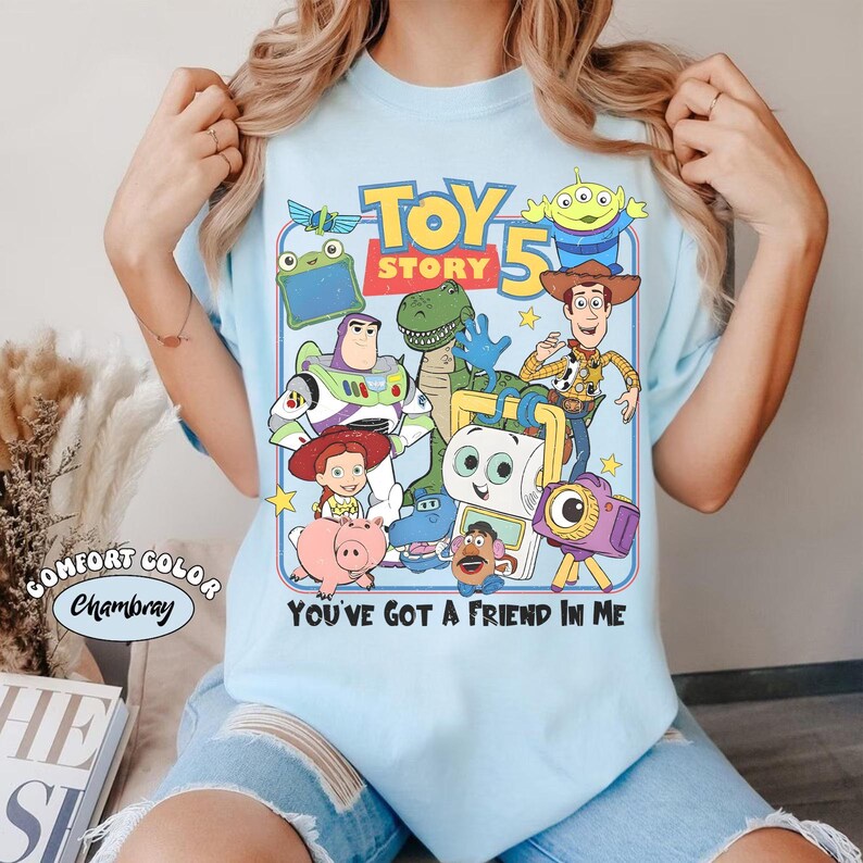 Pu&ograve; includere: T-shirt azzurra con una grafica colorata dei personaggi di Toy Story 5, tra cui Woody, Buzz Lightyear e Rex. Il design include anche il testo "TOY STORY 5" e la frase "YOU'VE GOT A FRIEND IN ME".