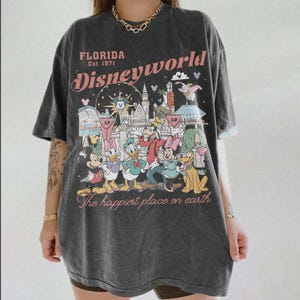 Peut inclure: T-shirt gris fonc&eacute; avec un graphisme color&eacute; des personnages et des monuments de Disney World. Le texte "Florida Est. 1971 Disneyworld" est imprim&eacute; au-dessus du graphisme, avec "The happiest place on earth" en dessous.