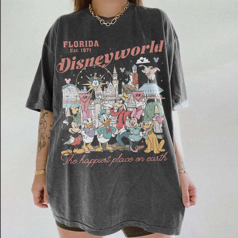 Peut inclure: T-shirt gris fonc&eacute; avec un graphisme color&eacute; des personnages et des monuments de Disney World. Le texte "Florida Est. 1971 Disneyworld" est imprim&eacute; au-dessus du graphisme, avec "The happiest place on earth" en dessous.