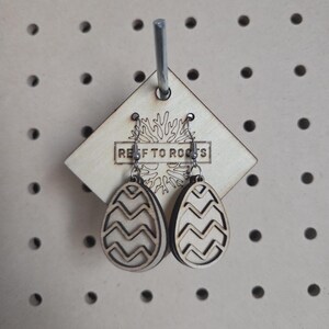 May include: Wooden Easter egg earrings with a zig-zag design, hanging from a wooden tag that reads "REEF TO ROOTS". The earrings are displayed on a pegboard.