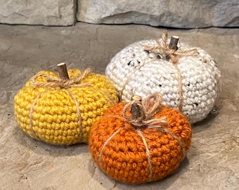 May include: Three crocheted pumpkins in shades of yellow, orange, and white. Each pumpkin has a small wooden stem and is wrapped with twine. The pumpkins are arranged on a stone surface, suggesting a fall or harvest theme.