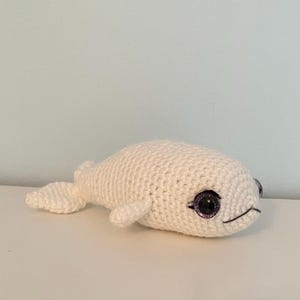 May include: A crocheted white whale plush toy with purple glitter eyes and a stitched smile. The handmade stuffed animal has fins and a tail. The whale is resting on a white surface.
