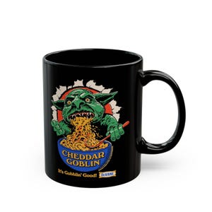 May include: Black ceramic mug featuring a green goblin graphic eating macaroni and cheese from a blue bowl. The text "Cheddar Goblin" and "It's Gobblin' Good!" are printed on the mug. A red spoon is in the bowl.