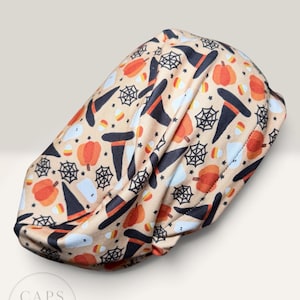 May include: A tan scrub cap featuring a repeating Halloween pattern of orange pumpkins, black witch hats, white ghosts, black spiderwebs, and candy corn. The fabric is gathered at the center, and a circular logo reads CAPS by CASEY.