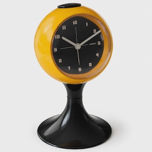 Könnte beinhalten: Eine kugelförmige gelbe Tischuhr mit schwarzem Zifferblatt und weißen Ziffern. Die Uhr hat dünne weiße Zeiger und das Wort ORBIT ist auf dem Zifferblatt aufgedruckt. Sie steht auf einem glänzend schwarzen Sockel mit einem breiten, runden Fuß.