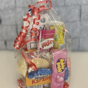 Könnte beinhalten: Ein in Zellophan verpackter Geschenkkorb mit verschiedenen Süßigkeiten und Snacks. Der Korb enthält eine rosa Hubba Bubba Kaugummi-Packung, eine Karam-Schachtel und andere bunte Leckereien. Ein rotes Band kringelt sich oben.