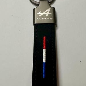 May include: Black leather keyring with a silver-coloured metal ring and a rectangular plate with the word "ALPINE" and the number "4". The keyring has a vertical stripe in red, white, and blue.