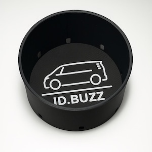 May include: Black circular car cup holder featuring a white van graphic and the text "ID.BUZZ". The interior has a textured pattern, and the sides have small ventilation holes. The cup holder is designed for a car.