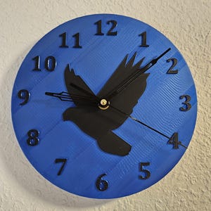 May include: A round blue wall clock with black numerals and hands. A black silhouette of a bird is in the center. The clock is mounted on a textured white wall.
