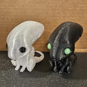May include: Two 3D-printed alien figurines. One is white with black eyes, the other is black with green eyes. Both have a similar design, with a large head, small body, and a tail. The figurines are sitting on a dark surface.
