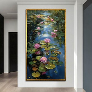 May include: A framed painting of a pond scene with pink water lilies and lily pads. The water reflects the sky and surrounding foliage. The painting is in a gold frame and hangs on a white wall.