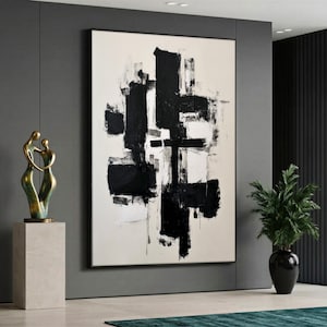 May include: Large abstract painting with black brushstrokes on a cream background, framed in black. A gold sculpture of two figures embracing sits on a stone pedestal. A black vase holds a green plant.