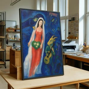 May include: A framed painting with a surrealist theme. The artwork features a woman in a red dress and veil, holding a green heart. A goat playing a guitar and figures in blue are also depicted against a blue backdrop. The painting is displayed on a wooden table.