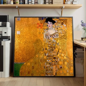 Puede incluir: Impresión enmarcada de una pintura de Gustav Klimt. La obra presenta a una mujer con un vestido dorado con patrones intrincados y un sombrero negro. El fondo es un mosaico de tonos dorados y amarillos, con una sección verde en la parte inferior.