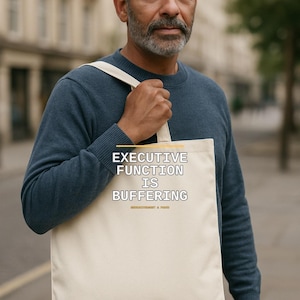 Op de afbeelding: Een beige canvas draagtas met de tekst "EXECUTIVE FUNCTION IS BUFFERING" in zwarte hoofdletters, een gele horizontale lijn boven de tekst en kleinere gele tekst eronder, allemaal gedrukt op de voorzijde van de tas.