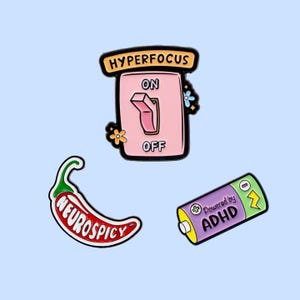 Op de afbeelding: Drie emaille pins op een lichtblauwe achtergrond. Eén pin is een roze schakelaar met de woorden "HYPERFOCUS", "ON" en "OFF". Een andere is een rode chilipeper met de woorden "NEUROSPICY". De laatste is een paarse batterij met "Powered by ADHD".