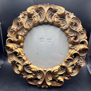 May include: An ornate, circular picture frame with a gold finish. The frame has detailed, swirling scrollwork around the edge. The centre has a grey backing with two small holes for photo placement. A decorative item for home decor.