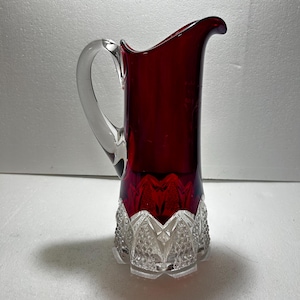 May include: A ruby red glass pitcher with a clear glass handle. The base of the pitcher features a cut crystal design. The pitcher is approximately 10 inches tall.