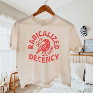 Könnte beinhalten: Cremefarbenes T-Shirt mit roter Grafik. Die Grafik zeigt einen Vogel, der einen Zweig hält, und die Worte "RADICALIZED BY BASIC DECENCY" in einem kreisförmigen Design. Das T-Shirt hängt an einem Holzbügel.
