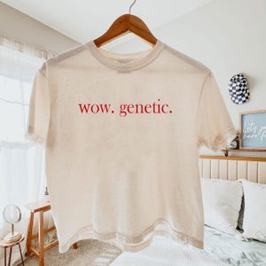 Peut inclure: Un t-shirt crème avec le texte rouge "wow. genetic.". Le t-shirt à manches courtes est suspendu à un cintre en bois. Le t-shirt est fait d'une matière douce et légère.