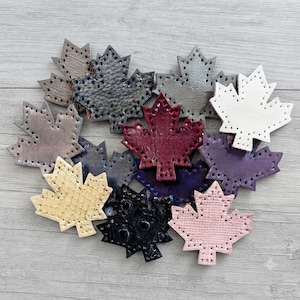 Puede incluir: Una colección de recortes con forma de hoja de arce en varios colores, incluyendo blanco, negro, rosa, amarillo, morado, burdeos y gris. Cada hoja presenta una superficie texturizada y una serie de pequeños agujeros espaciados uniformemente.