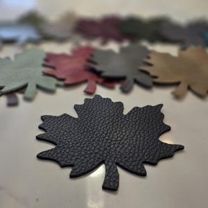 May include: A black leather maple leaf shape in focus, with a textured surface. Several other maple leaf shapes in various colors are blurred in the background, including green, red, and gold.
