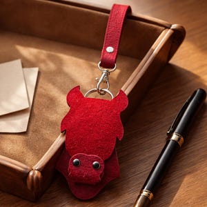 Peut inclure: Un porte-clés en feutre rouge en forme de taureau, avec un fermoir argenté et une lanière rouge assortie. Le taureau a deux yeux noirs en forme de boutons et une tête détaillée. Le porte-clés est posé sur une surface en bois avec un stylo et un plateau.