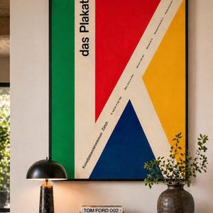 May include: A framed poster with a striking geometric design. The artwork features a green vertical stripe, a red section, a yellow triangle, and a blue triangle, with the words "das Plakat" in white. The poster is displayed above a black marble table.