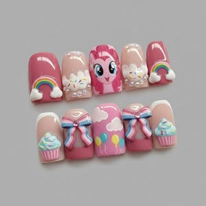 May include: A set of ten press-on nails with a pink and nude color scheme. Designs include a rainbow, a cartoon pony, sprinkles, bows, clouds, balloons, and cupcakes. The nails are decorated with 3D embellishments.