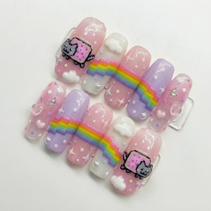 May include: A set of ten artificial nails with a whimsical design. The nails feature a pink base with various decorations, including rainbows, clouds, stars, and a cartoon cat. Some nails have a glittery finish, and others have pearl accents.