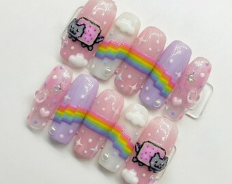 May include: A set of ten artificial nails with a whimsical design. The nails feature a pink base with various decorations, including rainbows, clouds, stars, and a cartoon cat. Some nails have a glittery finish, and others have pearl accents.