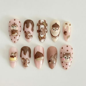 May include: A collection of ten almond-shaped false nails with autumnal designs. The nails showcase squirrels, acorns, and chocolate drizzles. The colour palette includes pink, brown, and cream, with some nails featuring a polka dot pattern.