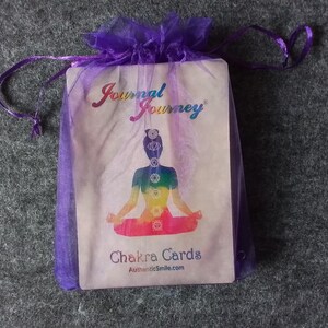 May include: A purple organza bag containing a deck of "Chakra Cards". The cards feature a colourful illustration of a person in a seated yoga pose with chakra symbols. The bag has a drawstring closure and the words "Journal Journey" are printed on the card.