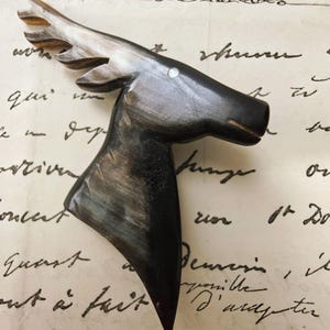 Pode incluir: Um broche esculpido em forma de perfil de cabeça de veado, feito de chifre escuro polido com estrias naturais cinzentas e pretas. O design apresenta um pequeno ponto branco incrustado para o olho e chifres detalhados em camadas.
