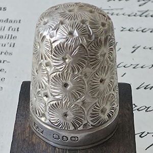 May include: A silver thimble featuring a repeating embossed floral pattern across its dome. The base of the thimble displays a smooth band with four distinct hallmarks stamped into the metal, indicating its origin and composition.