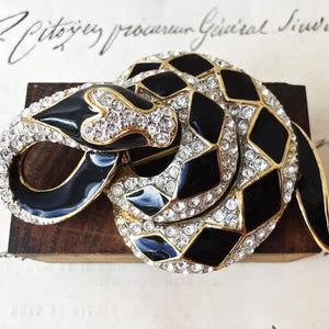 May include: A coiled snake brooch featuring a gold-tone metal frame with alternating black enamel diamond-shaped segments and clear sparkling rhinestones. The serpent is depicted in a curled position with its head resting on its body.