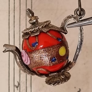 May include: A miniature teapot charm with a red glass body, adorned with gold, blue, pink, and yellow accents. The teapot has a silver-colored metal handle, spout, and lid, creating a vintage aesthetic. The charm is suspended from a silver loop.