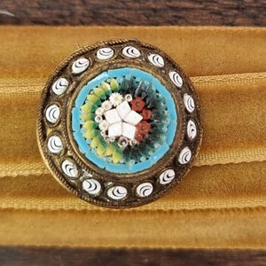 May include: A round, antique brooch with a mosaic design. The brooch features a central mosaic of white, red, and yellow flowers surrounded by a light blue border. The outer edge has a gold-colored metal frame with white and gold accents.